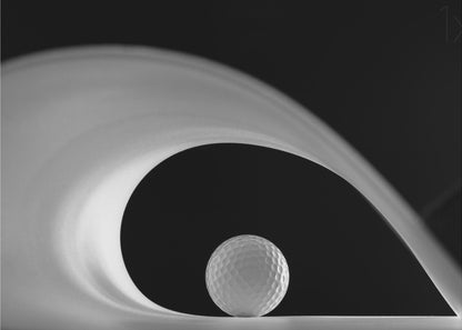 A dramatic black and white still life photo featuring a golf ball set against a stark black background. A large, abstract white shape swirls gracefully over the ball, creating a dynamic, wave-like composition. The image is presented within a silver frame. Decor