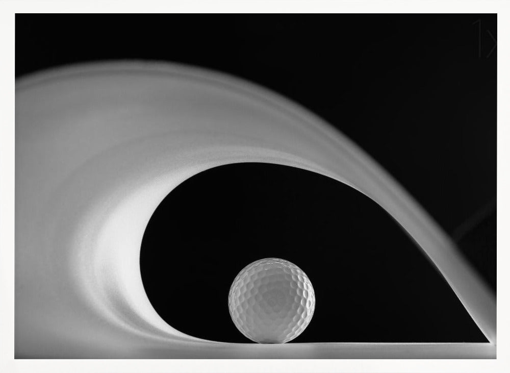 A dramatic black and white still life photo featuring a golf ball set against a stark black background. A large, abstract white shape swirls gracefully over the ball, creating a dynamic, wave-like composition. The image is presented within a silver frame. Decor