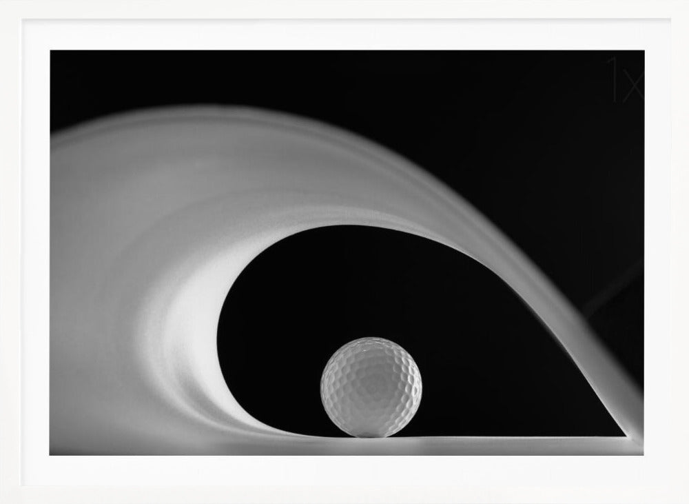 A dramatic black and white still life photo featuring a golf ball set against a stark black background. A large, abstract white shape swirls gracefully over the ball, creating a dynamic, wave-like composition. The image is presented within a silver frame. Decor