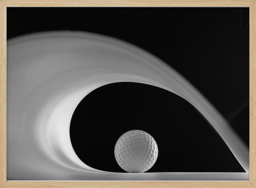 A dramatic black and white still life photo featuring a golf ball set against a stark black background. A large, abstract white shape swirls gracefully over the ball, creating a dynamic, wave-like composition. The image is presented within a silver frame. Decor
