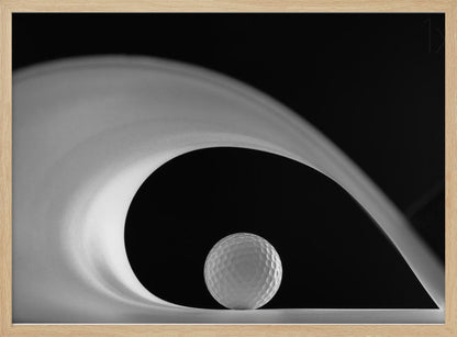 A dramatic black and white still life photo featuring a golf ball set against a stark black background. A large, abstract white shape swirls gracefully over the ball, creating a dynamic, wave-like composition. The image is presented within a silver frame. Decor