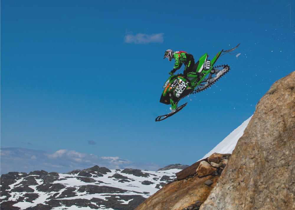 A dynamic action shot of a person in full gear launching a green snowmobile into the air off a rocky, snow-covered cliff, set against a backdrop of a vivid blue sky and distant snowy mountains. The image is enclosed in a silver frame. Wall Art