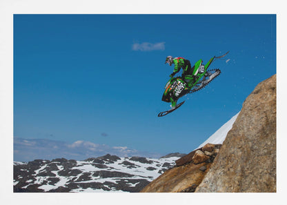 A dynamic action shot of a person in full gear launching a green snowmobile into the air off a rocky, snow-covered cliff, set against a backdrop of a vivid blue sky and distant snowy mountains. The image is enclosed in a silver frame. Wall Art