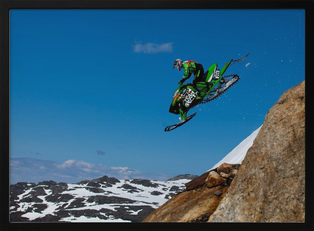 A dynamic action shot of a person in full gear launching a green snowmobile into the air off a rocky, snow-covered cliff, set against a backdrop of a vivid blue sky and distant snowy mountains. The image is enclosed in a silver frame. Wall Art