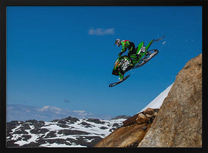 A dynamic action shot of a person in full gear launching a green snowmobile into the air off a rocky, snow-covered cliff, set against a backdrop of a vivid blue sky and distant snowy mountains. The image is enclosed in a silver frame. Wall Art