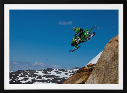 A dynamic action shot of a person in full gear launching a green snowmobile into the air off a rocky, snow-covered cliff, set against a backdrop of a vivid blue sky and distant snowy mountains. The image is enclosed in a silver frame. Wall Art