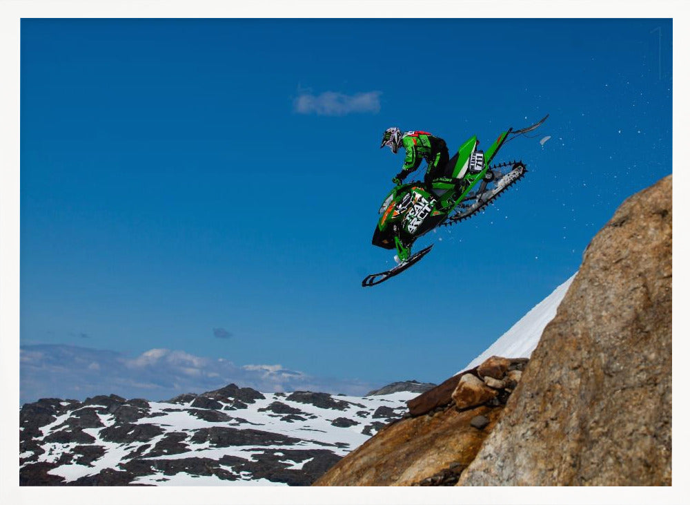 A dynamic action shot of a person in full gear launching a green snowmobile into the air off a rocky, snow-covered cliff, set against a backdrop of a vivid blue sky and distant snowy mountains. The image is enclosed in a silver frame. Wall Art