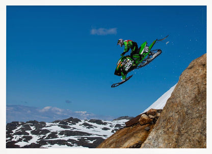 A dynamic action shot of a person in full gear launching a green snowmobile into the air off a rocky, snow-covered cliff, set against a backdrop of a vivid blue sky and distant snowy mountains. The image is enclosed in a silver frame. Wall Art