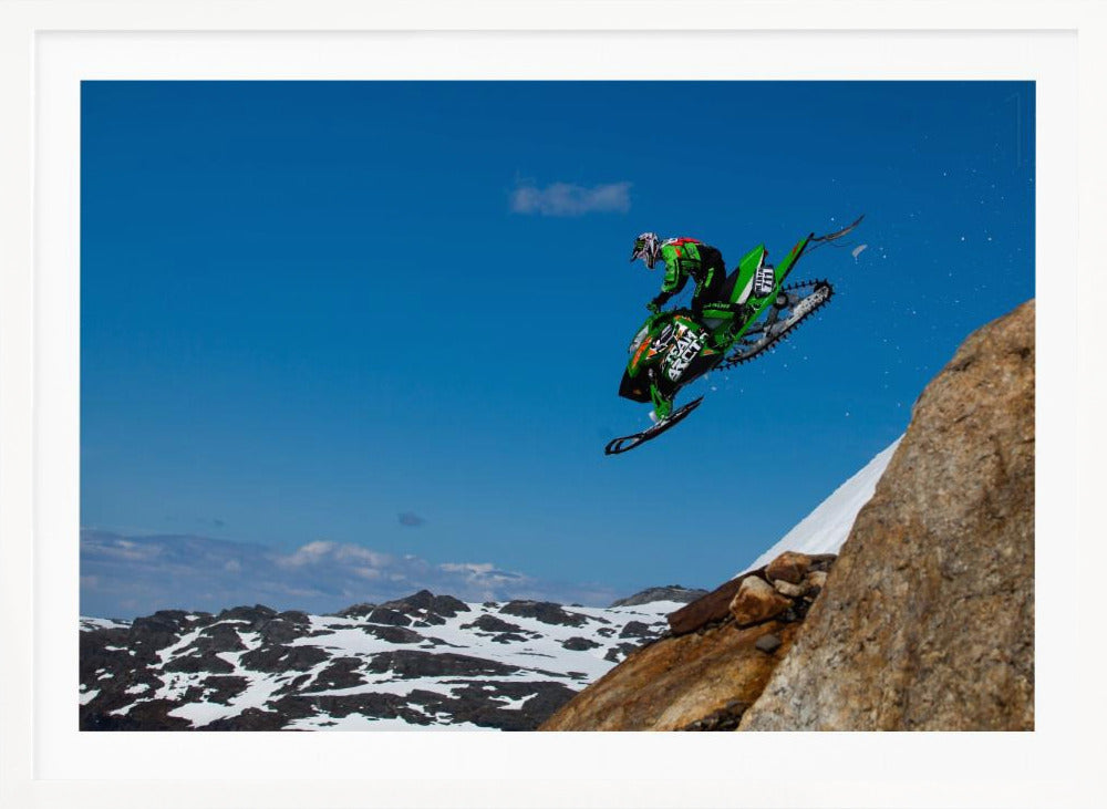 A dynamic action shot of a person in full gear launching a green snowmobile into the air off a rocky, snow-covered cliff, set against a backdrop of a vivid blue sky and distant snowy mountains. The image is enclosed in a silver frame. Wall Art
