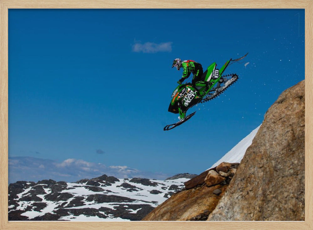 A dynamic action shot of a person in full gear launching a green snowmobile into the air off a rocky, snow-covered cliff, set against a backdrop of a vivid blue sky and distant snowy mountains. The image is enclosed in a silver frame. Wall Art
