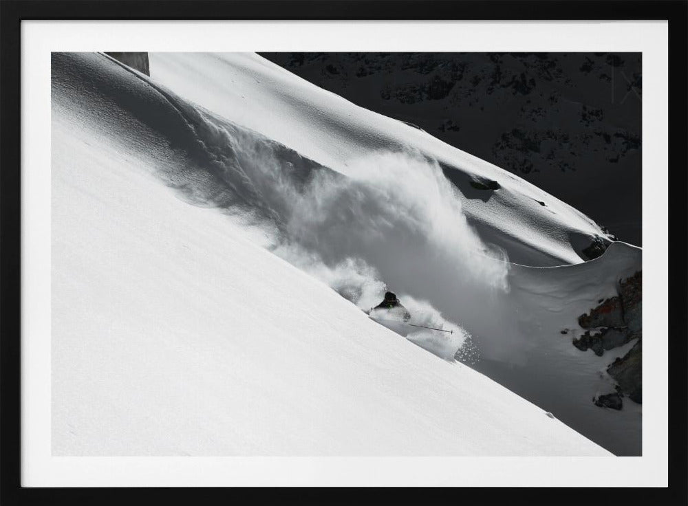 A dramatic black and white photograph of a skier speeding down a steep, snow-covered mountain, kicking up a large cloud of powder. The image is framed with a brushed silver border. Decor