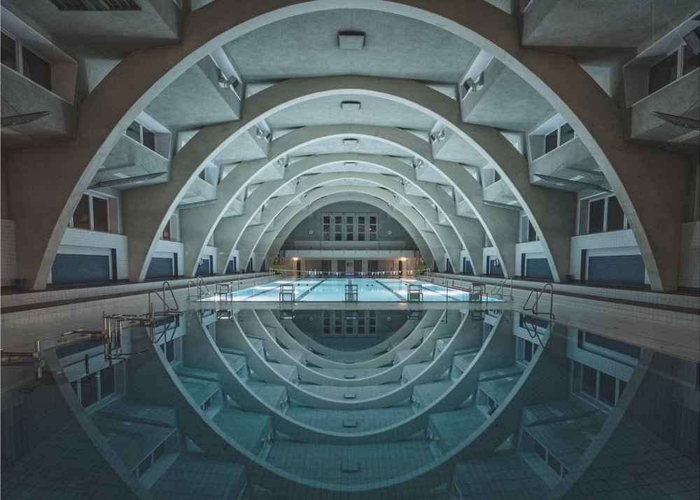 An atmospheric photograph of an empty indoor swimming pool with remarkable Brutalist-style architecture. A series of large, repeating concrete arches span the length of the pool, creating a tunnel-like effect. The arches and lights are perfectly reflected in the still, blue water, forming a symmetrical, eye-like shape. Decor