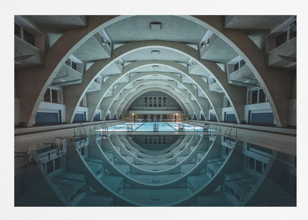 An atmospheric photograph of an empty indoor swimming pool with remarkable Brutalist-style architecture. A series of large, repeating concrete arches span the length of the pool, creating a tunnel-like effect. The arches and lights are perfectly reflected in the still, blue water, forming a symmetrical, eye-like shape. Decor