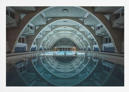 An atmospheric photograph of an empty indoor swimming pool with remarkable Brutalist-style architecture. A series of large, repeating concrete arches span the length of the pool, creating a tunnel-like effect. The arches and lights are perfectly reflected in the still, blue water, forming a symmetrical, eye-like shape. Decor
