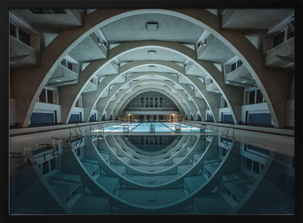 An atmospheric photograph of an empty indoor swimming pool with remarkable Brutalist-style architecture. A series of large, repeating concrete arches span the length of the pool, creating a tunnel-like effect. The arches and lights are perfectly reflected in the still, blue water, forming a symmetrical, eye-like shape. Decor