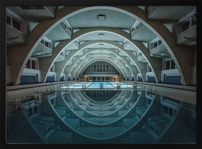 An atmospheric photograph of an empty indoor swimming pool with remarkable Brutalist-style architecture. A series of large, repeating concrete arches span the length of the pool, creating a tunnel-like effect. The arches and lights are perfectly reflected in the still, blue water, forming a symmetrical, eye-like shape. Decor