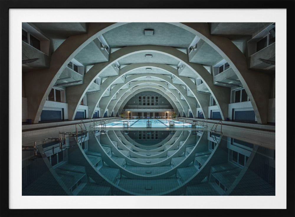 An atmospheric photograph of an empty indoor swimming pool with remarkable Brutalist-style architecture. A series of large, repeating concrete arches span the length of the pool, creating a tunnel-like effect. The arches and lights are perfectly reflected in the still, blue water, forming a symmetrical, eye-like shape. Decor