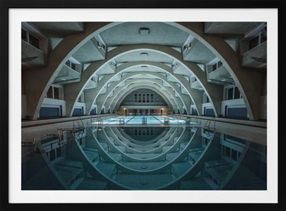 An atmospheric photograph of an empty indoor swimming pool with remarkable Brutalist-style architecture. A series of large, repeating concrete arches span the length of the pool, creating a tunnel-like effect. The arches and lights are perfectly reflected in the still, blue water, forming a symmetrical, eye-like shape. Decor