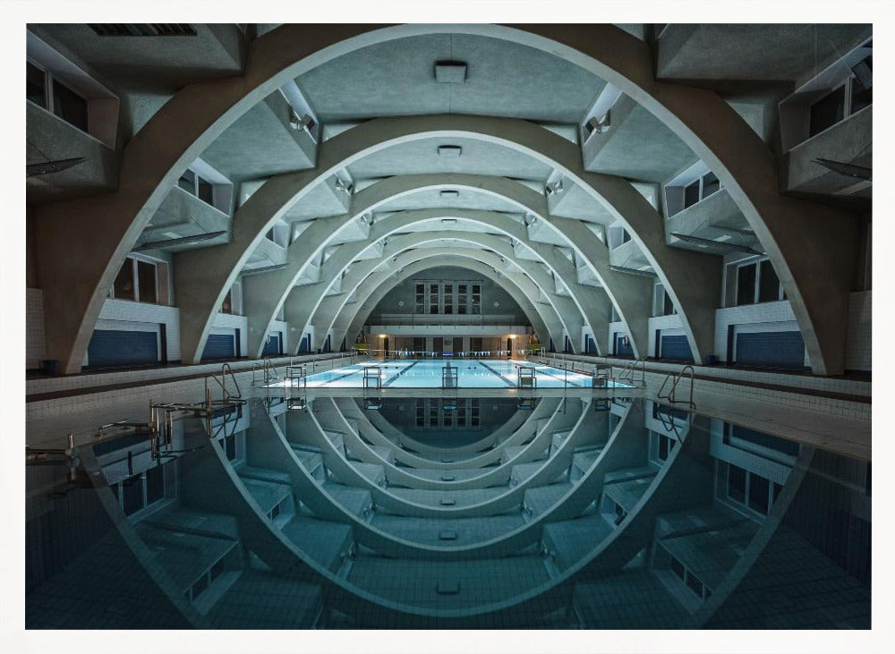 An atmospheric photograph of an empty indoor swimming pool with remarkable Brutalist-style architecture. A series of large, repeating concrete arches span the length of the pool, creating a tunnel-like effect. The arches and lights are perfectly reflected in the still, blue water, forming a symmetrical, eye-like shape. Decor