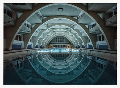 An atmospheric photograph of an empty indoor swimming pool with remarkable Brutalist-style architecture. A series of large, repeating concrete arches span the length of the pool, creating a tunnel-like effect. The arches and lights are perfectly reflected in the still, blue water, forming a symmetrical, eye-like shape. Decor