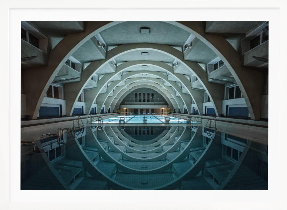 An atmospheric photograph of an empty indoor swimming pool with remarkable Brutalist-style architecture. A series of large, repeating concrete arches span the length of the pool, creating a tunnel-like effect. The arches and lights are perfectly reflected in the still, blue water, forming a symmetrical, eye-like shape. Decor