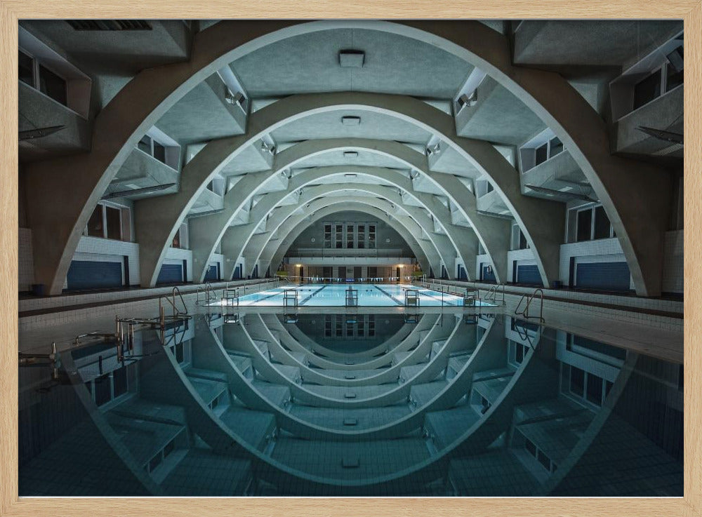 An atmospheric photograph of an empty indoor swimming pool with remarkable Brutalist-style architecture. A series of large, repeating concrete arches span the length of the pool, creating a tunnel-like effect. The arches and lights are perfectly reflected in the still, blue water, forming a symmetrical, eye-like shape. Decor