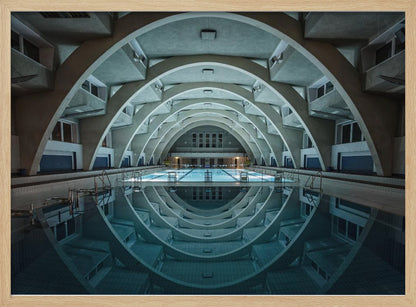 An atmospheric photograph of an empty indoor swimming pool with remarkable Brutalist-style architecture. A series of large, repeating concrete arches span the length of the pool, creating a tunnel-like effect. The arches and lights are perfectly reflected in the still, blue water, forming a symmetrical, eye-like shape. Decor