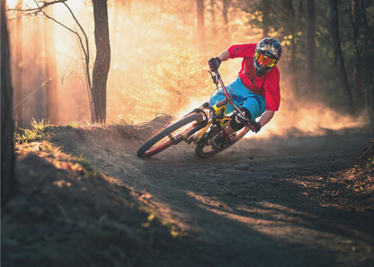 A mountain biker in a red shirt, blue shorts, and full-face helmet leans sharply into a dusty turn on a dirt trail in a forest. The setting sun casts a warm, golden light through the trees, creating a dramatic, backlit effect. Wall Art