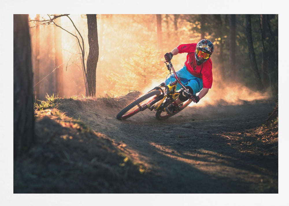 A mountain biker in a red shirt, blue shorts, and full-face helmet leans sharply into a dusty turn on a dirt trail in a forest. The setting sun casts a warm, golden light through the trees, creating a dramatic, backlit effect. Wall Art