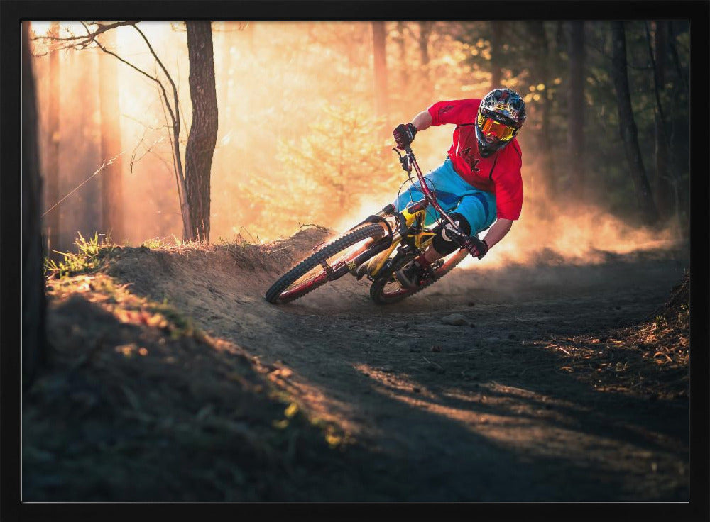 A mountain biker in a red shirt, blue shorts, and full-face helmet leans sharply into a dusty turn on a dirt trail in a forest. The setting sun casts a warm, golden light through the trees, creating a dramatic, backlit effect. Wall Art