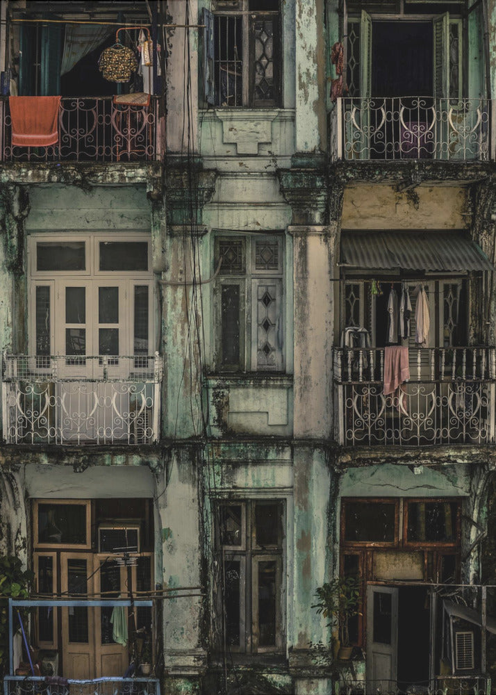 A tightly framed, symmetrical photograph of a decaying multi-story apartment building facade. The pale green paint is peeling and stained with dark grime, revealing the building's age. Each of the nine visible sections features a balcony with ornate ironwork or a window, showing glimpses of daily life through laundry hanging out to dry and dimly lit interiors. Poster