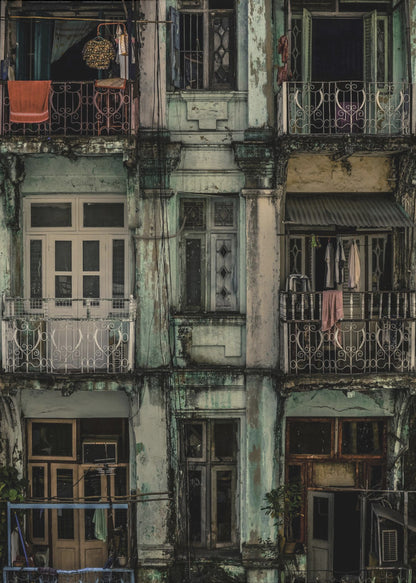 A tightly framed, symmetrical photograph of a decaying multi-story apartment building facade. The pale green paint is peeling and stained with dark grime, revealing the building's age. Each of the nine visible sections features a balcony with ornate ironwork or a window, showing glimpses of daily life through laundry hanging out to dry and dimly lit interiors. Poster