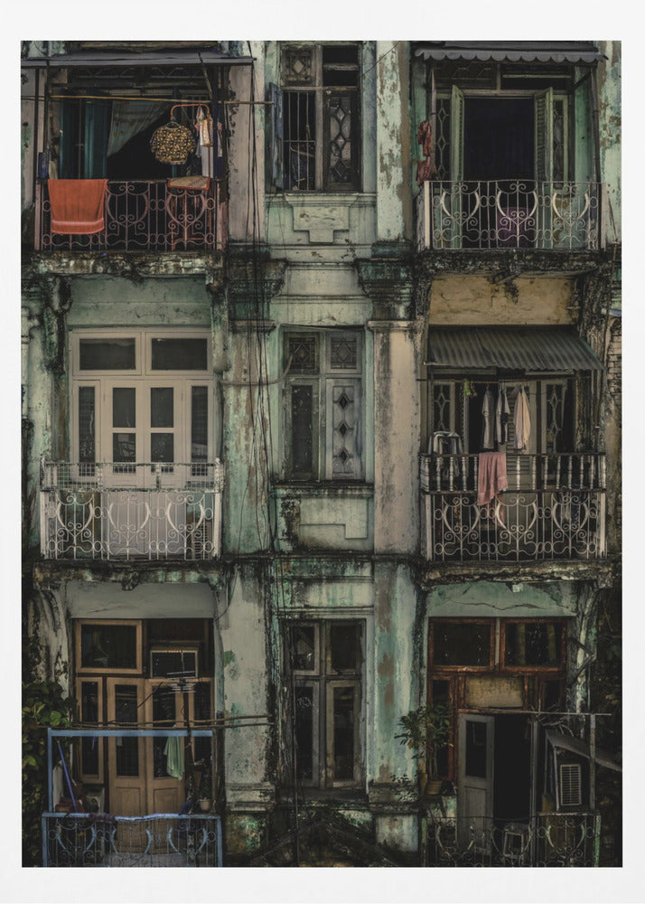 A tightly framed, symmetrical photograph of a decaying multi-story apartment building facade. The pale green paint is peeling and stained with dark grime, revealing the building's age. Each of the nine visible sections features a balcony with ornate ironwork or a window, showing glimpses of daily life through laundry hanging out to dry and dimly lit interiors. Poster