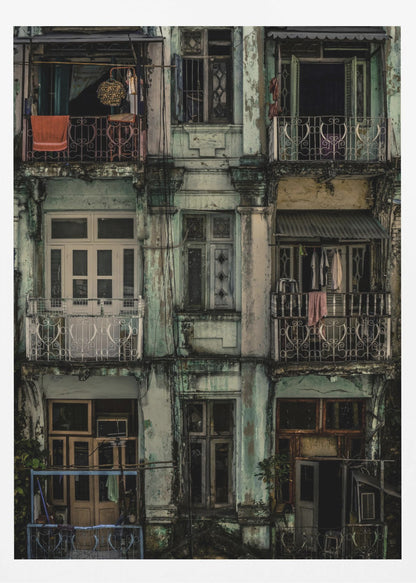 A tightly framed, symmetrical photograph of a decaying multi-story apartment building facade. The pale green paint is peeling and stained with dark grime, revealing the building's age. Each of the nine visible sections features a balcony with ornate ironwork or a window, showing glimpses of daily life through laundry hanging out to dry and dimly lit interiors. Poster