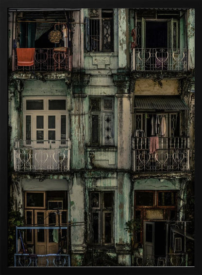 A tightly framed, symmetrical photograph of a decaying multi-story apartment building facade. The pale green paint is peeling and stained with dark grime, revealing the building's age. Each of the nine visible sections features a balcony with ornate ironwork or a window, showing glimpses of daily life through laundry hanging out to dry and dimly lit interiors. Poster