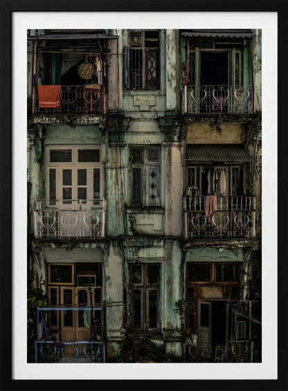 A tightly framed, symmetrical photograph of a decaying multi-story apartment building facade. The pale green paint is peeling and stained with dark grime, revealing the building's age. Each of the nine visible sections features a balcony with ornate ironwork or a window, showing glimpses of daily life through laundry hanging out to dry and dimly lit interiors. Poster
