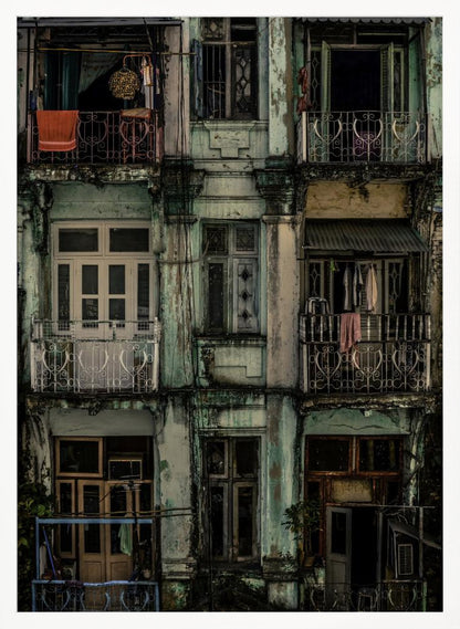 A tightly framed, symmetrical photograph of a decaying multi-story apartment building facade. The pale green paint is peeling and stained with dark grime, revealing the building's age. Each of the nine visible sections features a balcony with ornate ironwork or a window, showing glimpses of daily life through laundry hanging out to dry and dimly lit interiors. Poster