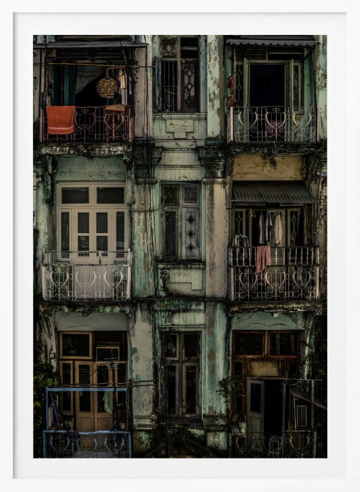 A tightly framed, symmetrical photograph of a decaying multi-story apartment building facade. The pale green paint is peeling and stained with dark grime, revealing the building's age. Each of the nine visible sections features a balcony with ornate ironwork or a window, showing glimpses of daily life through laundry hanging out to dry and dimly lit interiors. Poster