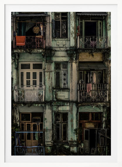 A tightly framed, symmetrical photograph of a decaying multi-story apartment building facade. The pale green paint is peeling and stained with dark grime, revealing the building's age. Each of the nine visible sections features a balcony with ornate ironwork or a window, showing glimpses of daily life through laundry hanging out to dry and dimly lit interiors. Poster