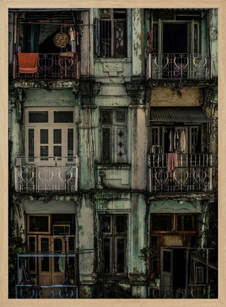 A tightly framed, symmetrical photograph of a decaying multi-story apartment building facade. The pale green paint is peeling and stained with dark grime, revealing the building's age. Each of the nine visible sections features a balcony with ornate ironwork or a window, showing glimpses of daily life through laundry hanging out to dry and dimly lit interiors. Poster