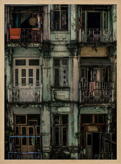 A tightly framed, symmetrical photograph of a decaying multi-story apartment building facade. The pale green paint is peeling and stained with dark grime, revealing the building's age. Each of the nine visible sections features a balcony with ornate ironwork or a window, showing glimpses of daily life through laundry hanging out to dry and dimly lit interiors. Poster