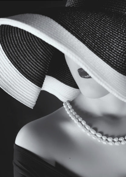 A glamorous black and white close-up photo of a woman wearing a large, striped sun hat that hides her eyes, revealing only her dark lips. She wears a pearl necklace and an off-the-shoulder top against a dark background, creating a mysterious and elegant mood. Wall Art