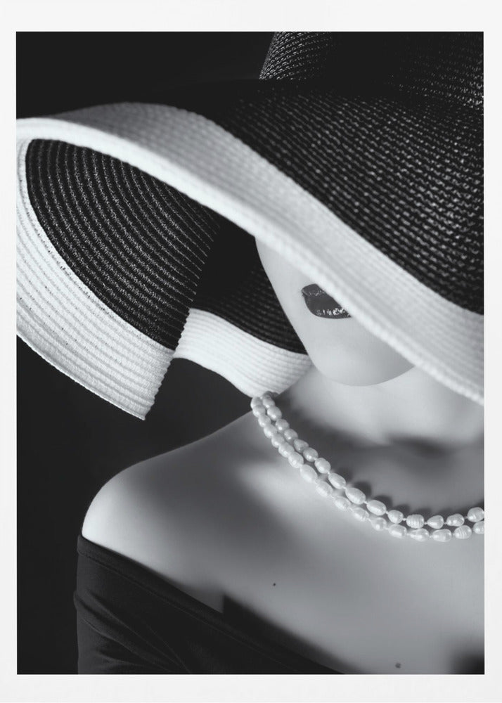 A glamorous black and white close-up photo of a woman wearing a large, striped sun hat that hides her eyes, revealing only her dark lips. She wears a pearl necklace and an off-the-shoulder top against a dark background, creating a mysterious and elegant mood. Wall Art