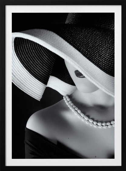 A glamorous black and white close-up photo of a woman wearing a large, striped sun hat that hides her eyes, revealing only her dark lips. She wears a pearl necklace and an off-the-shoulder top against a dark background, creating a mysterious and elegant mood. Wall Art