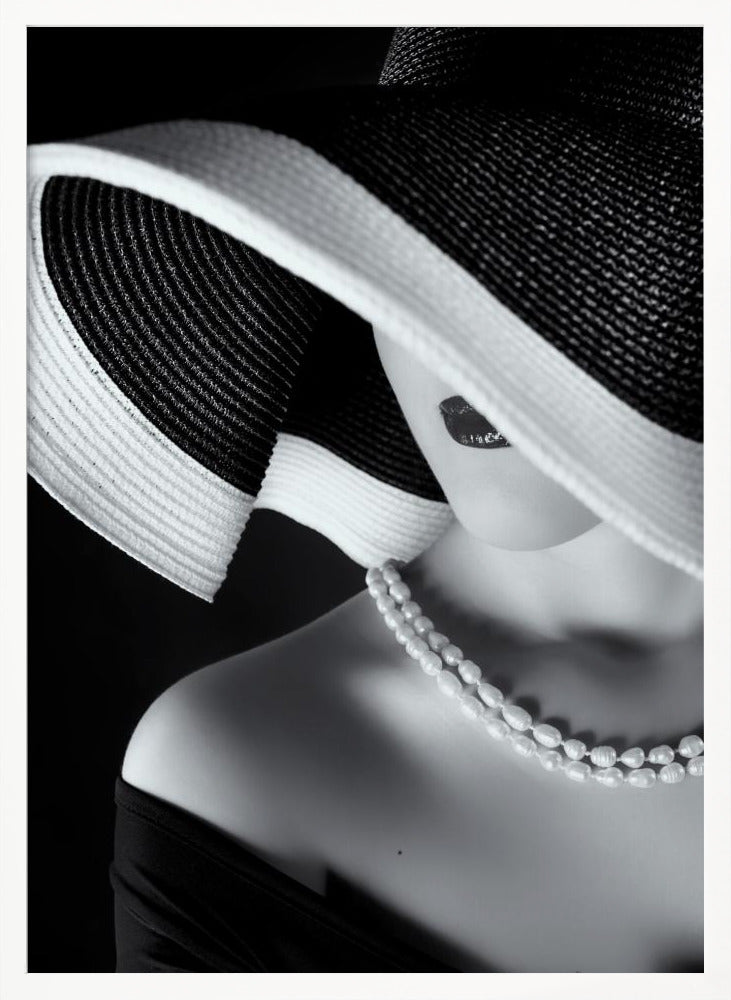 A glamorous black and white close-up photo of a woman wearing a large, striped sun hat that hides her eyes, revealing only her dark lips. She wears a pearl necklace and an off-the-shoulder top against a dark background, creating a mysterious and elegant mood. Wall Art
