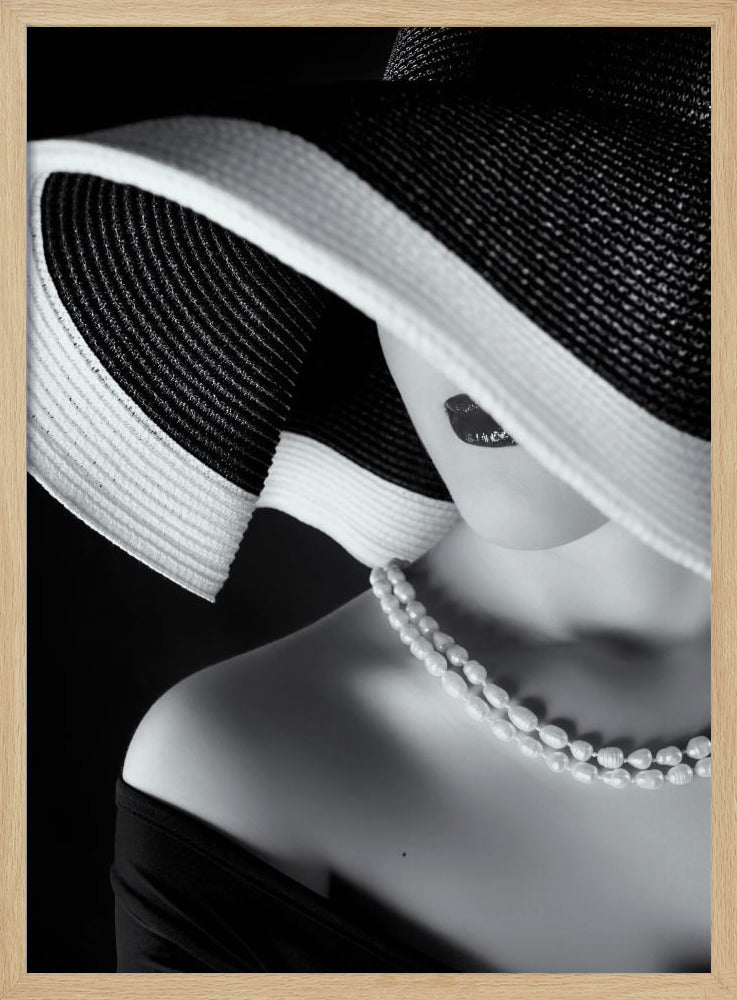 A glamorous black and white close-up photo of a woman wearing a large, striped sun hat that hides her eyes, revealing only her dark lips. She wears a pearl necklace and an off-the-shoulder top against a dark background, creating a mysterious and elegant mood. Wall Art