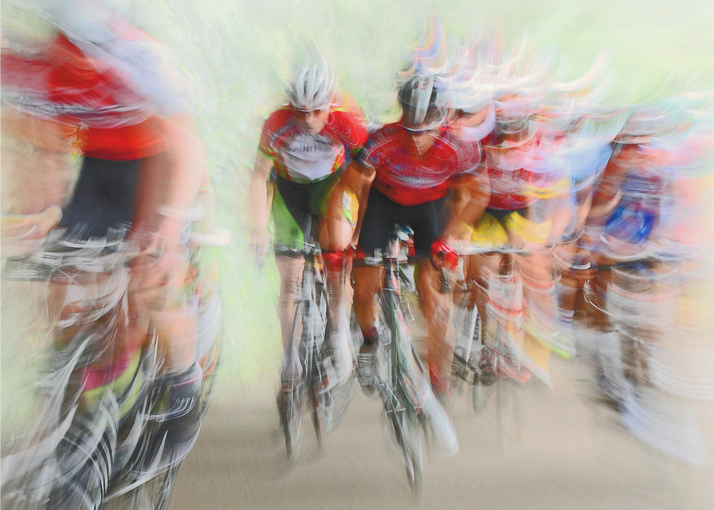An abstract photograph of a group of cyclists in a race, captured with motion blur to create a dynamic sense of speed. The riders' red jerseys and the green background are streaked together, with a silver frame surrounding the image. Artwork