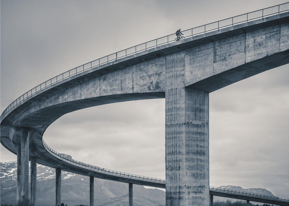 A black and white photograph of a lone cyclist riding across the top level of a massive, curving concrete bridge, which sweeps across the frame. In the background, a lower level of the bridge and distant mountains are visible under a cloudy sky. Decor