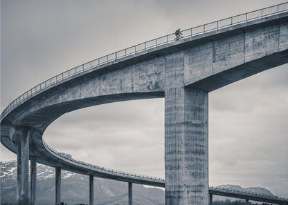 A black and white photograph of a lone cyclist riding across the top level of a massive, curving concrete bridge, which sweeps across the frame. In the background, a lower level of the bridge and distant mountains are visible under a cloudy sky. Decor