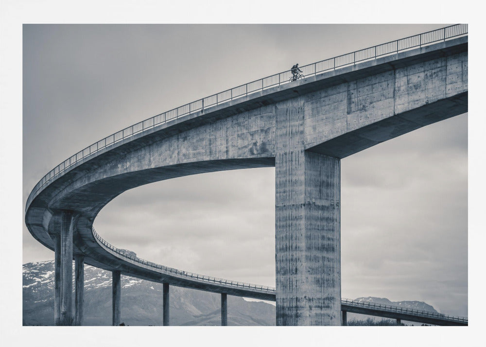 A black and white photograph of a lone cyclist riding across the top level of a massive, curving concrete bridge, which sweeps across the frame. In the background, a lower level of the bridge and distant mountains are visible under a cloudy sky. Decor
